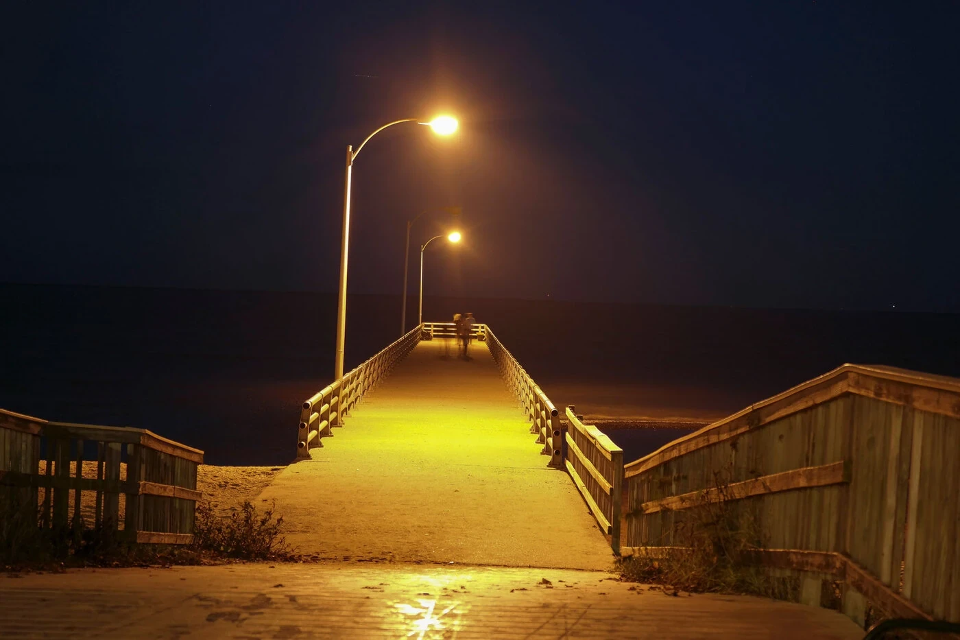 A lamplit boardwalk leading toward dark water at night.